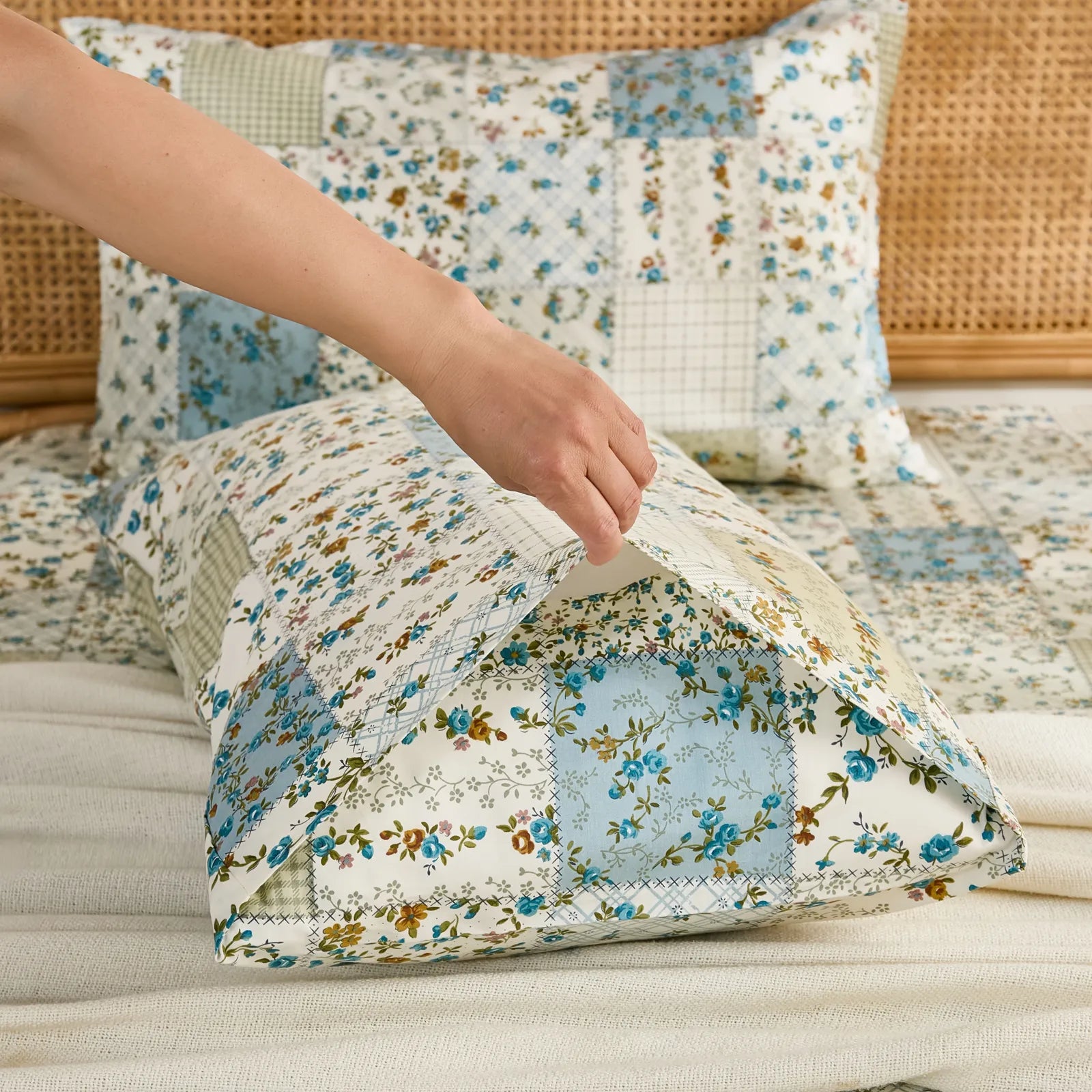 A hand tucking a pillow into the cotton pillowcase, showing the envelope closure and blue floral patchwork pattern.