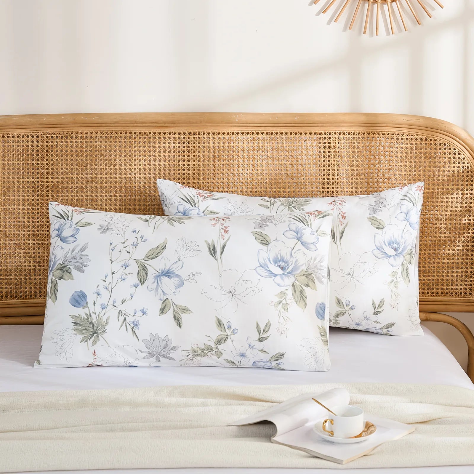 A pair of white cotton pillowcases with a blue and grey watercolor floral print, styled against a rattan headboard.