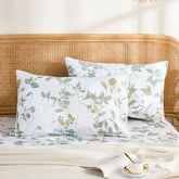 A pair of white cotton pillowcases with a fresh green botanical leaf and vine print, styled on a bed with a rattan headboard.