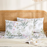 A pair of white cotton pillowcases with a lush green and pink tropical jungle print, styled against a rattan headboard.