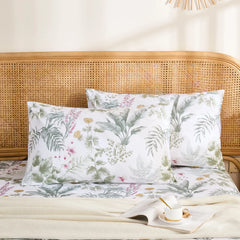 A pair of white cotton pillowcases with a lush green and pink tropical jungle print, styled against a rattan headboard.