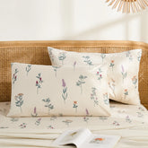 A pair of cream cotton pillowcases with purple and sage wildflower print displayed on a bed with a rattan headboard.
