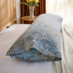 An angled shot of a blue paisley print cotton body pillow resting on a white bed, highlighting the rich pattern and fabric drape near a window.