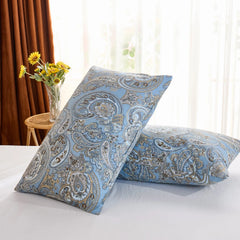 An angled shot of two light blue paisley print cotton pillowcases leaning against each other on a white bed, with a vase of sunflowers in the background.