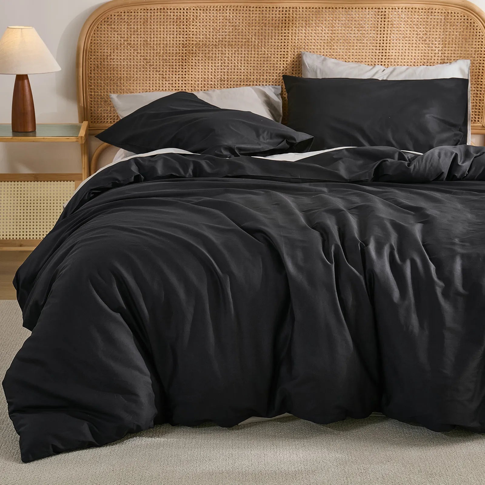 Angled close-up of black duvet showing natural folds, wooden lamp on woven nightstand with soft lighting.