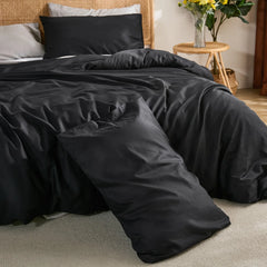 Front view of black cotton duvet with grey and black pillows against a woven rattan headboard, yellow flowers on side table.