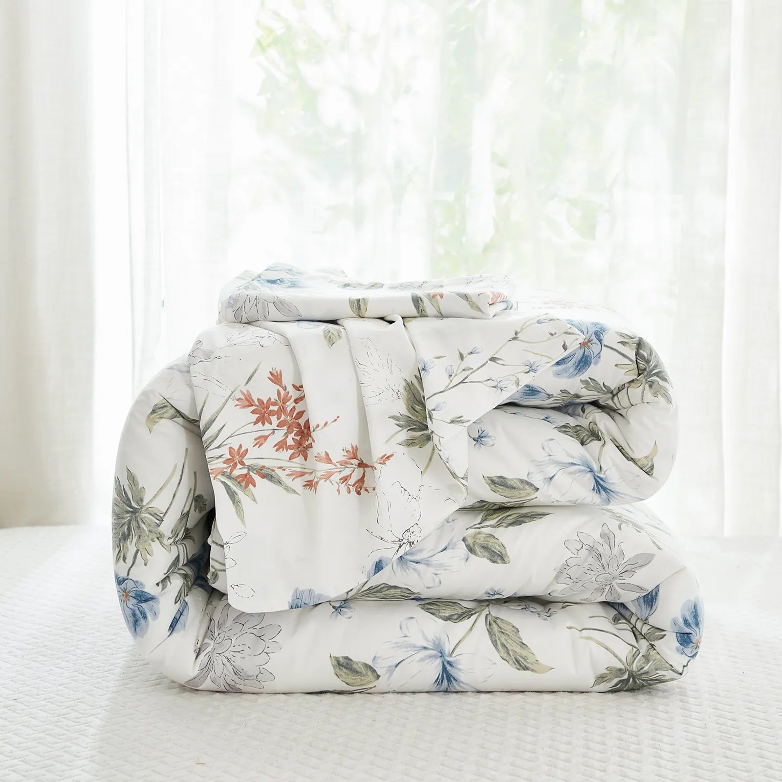 A white floral comforter set folded and stacked on a bed with a window in the background.