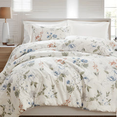 Angled view of a bed with a white floral comforter set, light wood headboard, and a bedside lamp.