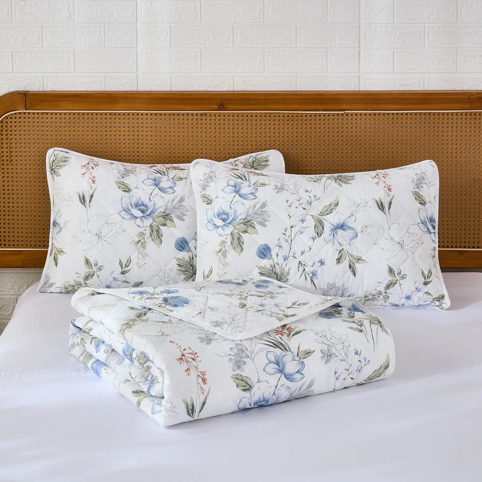 A neatly folded white and blue floral quilt set with two pillows on a bed with a rattan headboard.