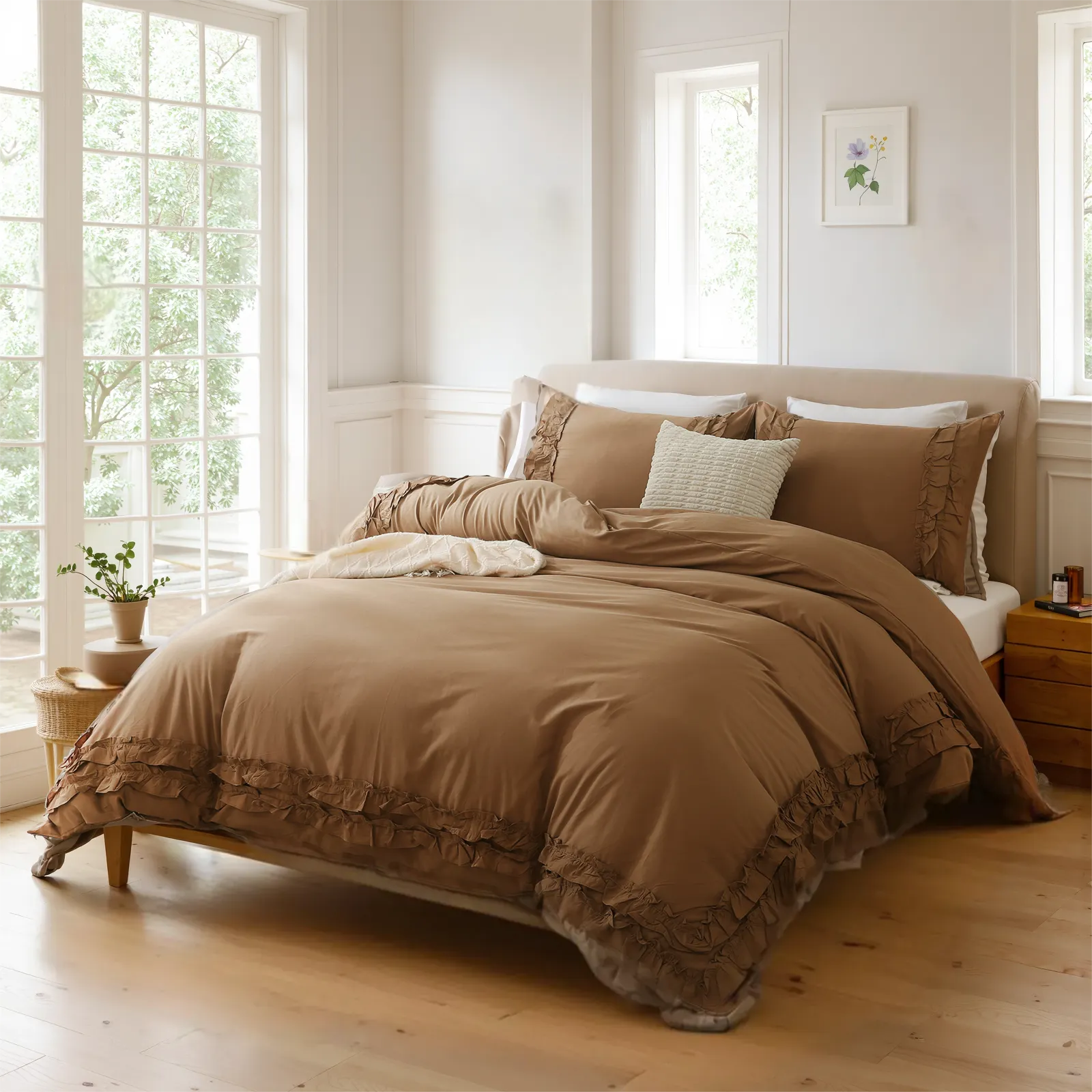 Brown cotton double ruffle bedding on a bed between wooden nightstands, under floral art and three large windows.
