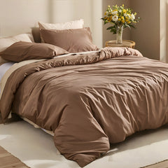 Angled close-up of brown duvet with white pillows and yellow-white flowers on round rattan side table.