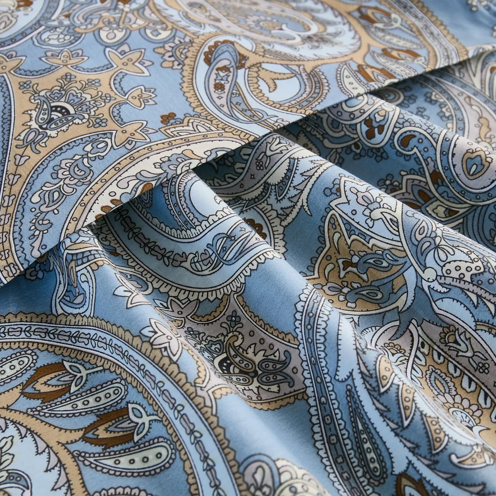 A close-up of the folded light blue cotton fabric, showcasing the detailed and intricate paisley pattern with brown and white accents.