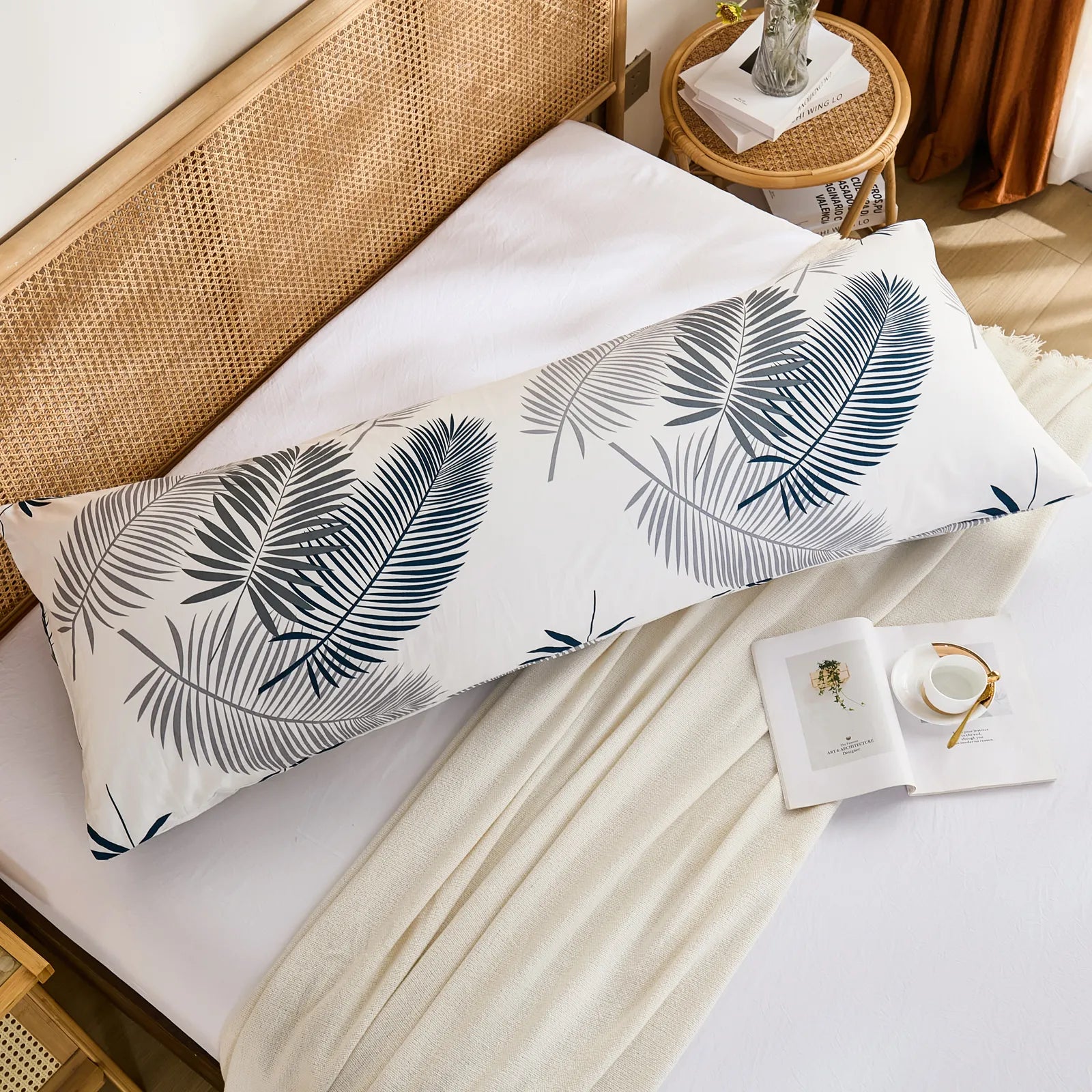 An overhead flat-lay shot of a cotton body pillowcase with a modern leaf print, placed on a white fitted sheet with a beige throw blanket.