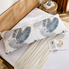 An overhead flat-lay shot of a cotton body pillowcase with a modern leaf print, placed on a white fitted sheet with a beige throw blanket.