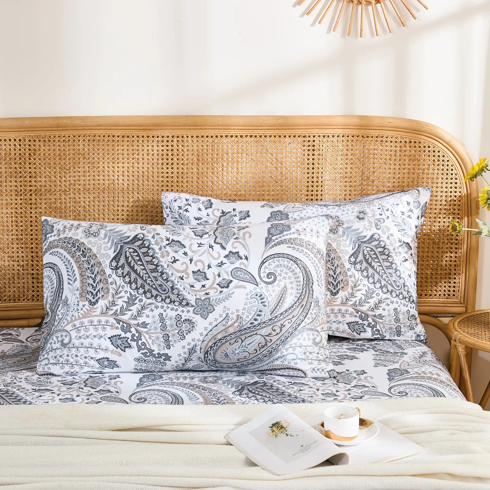 A pair of cotton grey paisley pillowcases arranged neatly on a bed with a rattan headboard, a vase of sunflowers, and an open book.