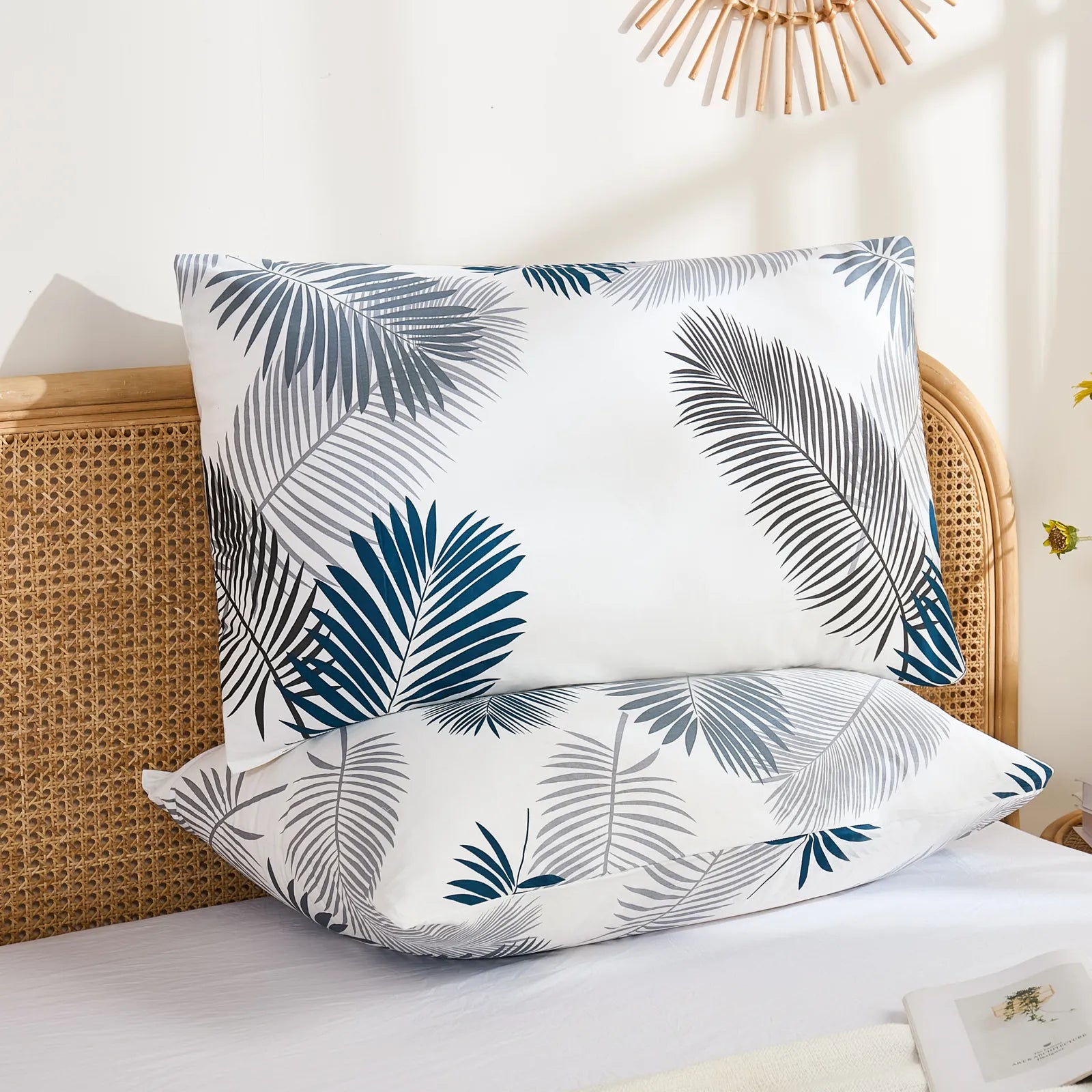A pair of cotton leaf print pillowcases are stacked on a bed with a rattan headboard, showing the full pattern and how they complement the bedroom decor.