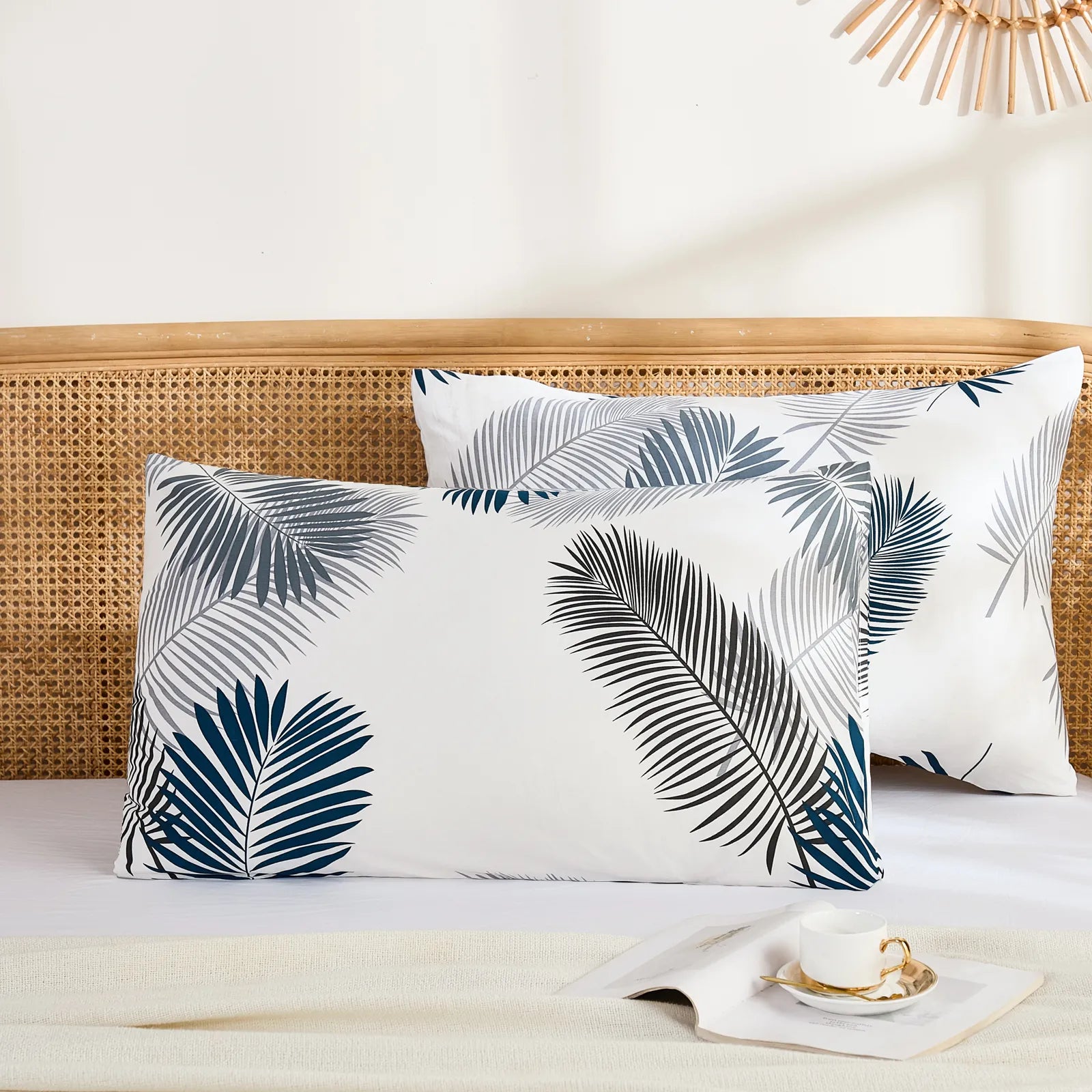 A wide view of two cotton pillowcases with a tropical leaf pattern, placed on a bed with a wicker headboard, a book, and a teacup.