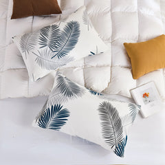 A flat-lay photo of two cotton leaf print pillowcases on a white quilted duvet, surrounded by brown and gold accent pillows.