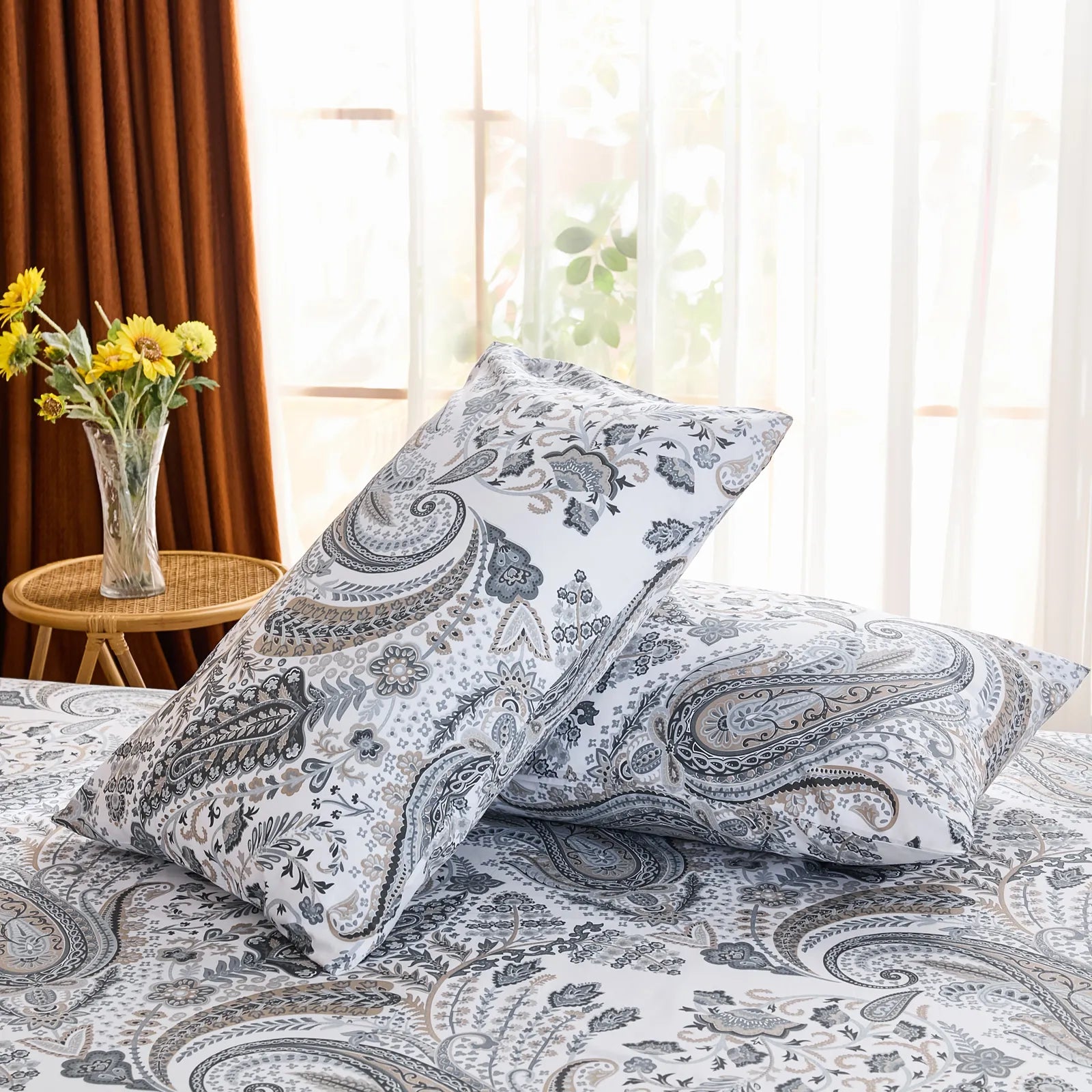 An angled view of two cotton paisley pillowcases propped up on a bed, with a large window and brown curtains providing a bright backdrop.