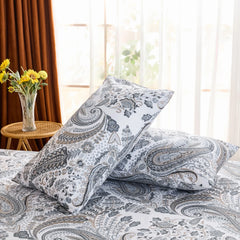 An angled view of two cotton paisley pillowcases propped up on a bed, with a large window and brown curtains providing a bright backdrop.
