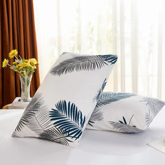 Two cotton pillowcases with a tropical leaf pattern are propped on a bed, with a large window and yellow sunflowers in the background.