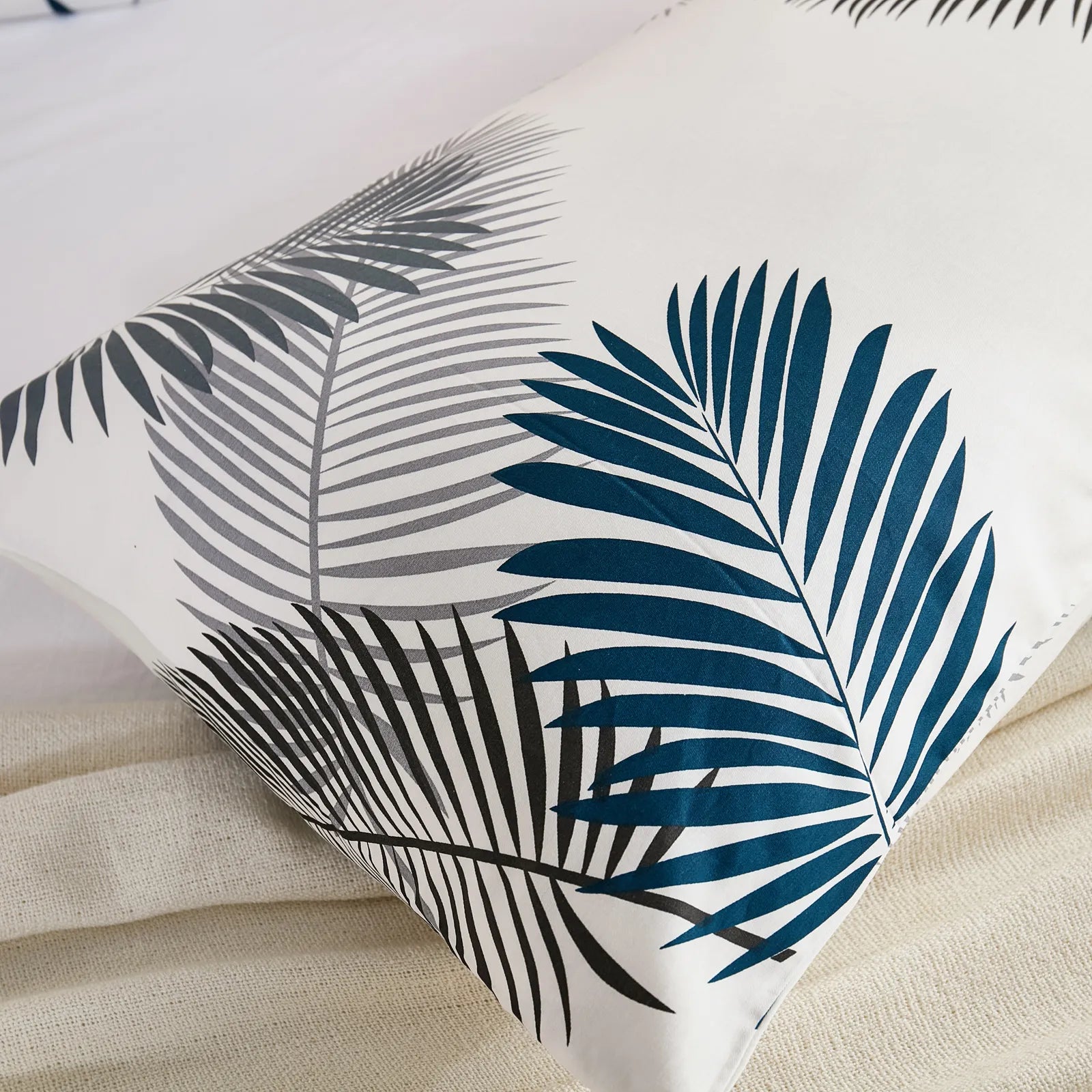 Two cotton pillowcases with a tropical leaf pattern in blue and gray, propped up against each other on a bed with a rattan headboard and sunlight streaming in.