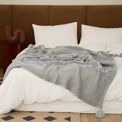 Light-grey knit throw blanket draped on a white bed with tassels, showing cable and open-knit textures.