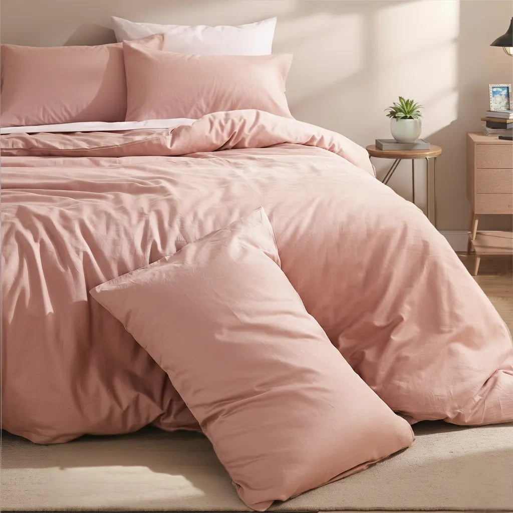 Dusty pink solid cotton duvet and pillows on a bed next to a round side table with a green plant and a wooden cabinet.