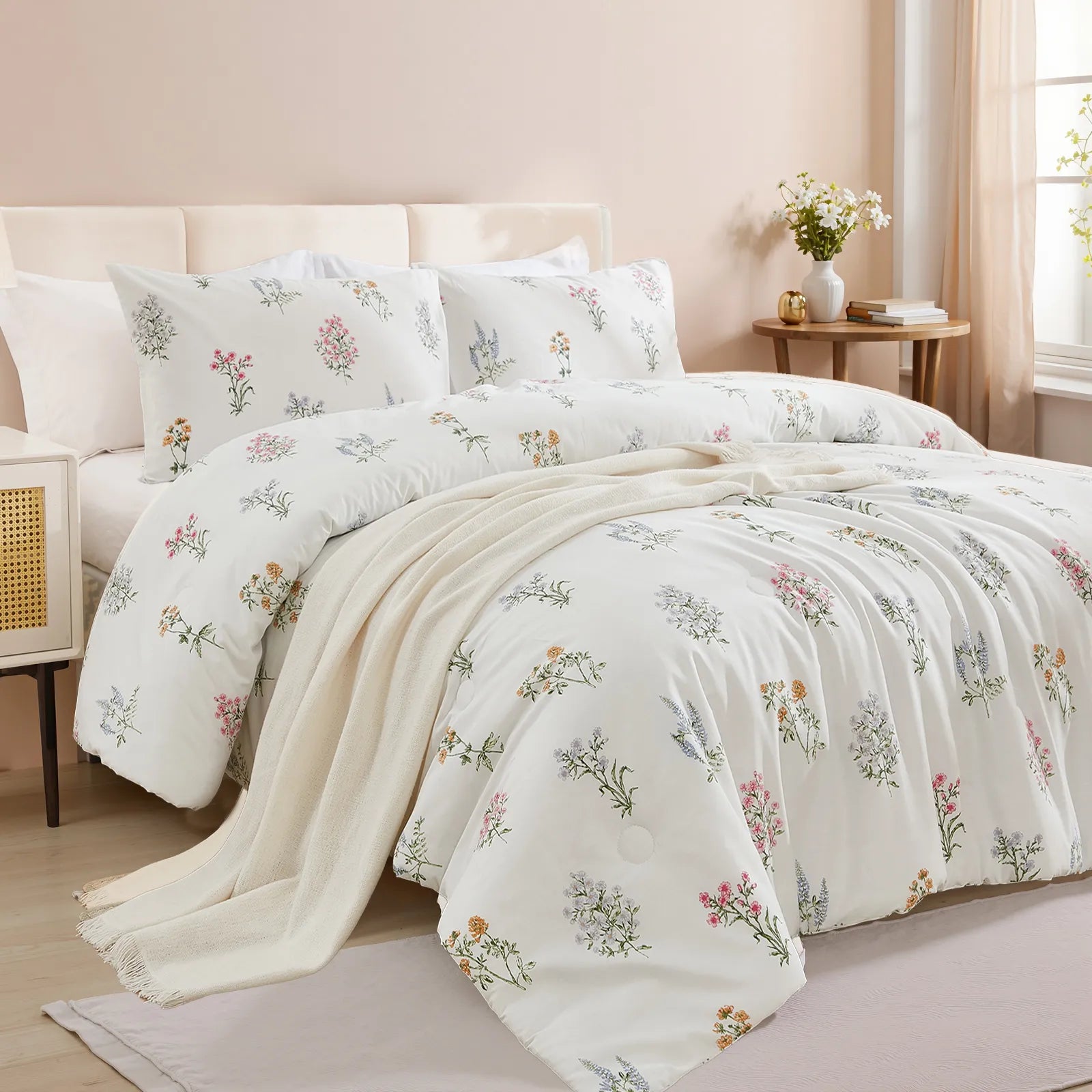 Angled view of a bed with a white comforter set featuring a pastel floral design, a beige blanket, and a light wood nightstand.
