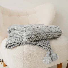 Folded light-grey cable-knit throw blanket with tassels neatly placed on a cream armchair.