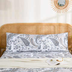 A long grey paisley cotton pillowcase is displayed on a bed with a rattan headboard, styled with a light-colored throw blanket.