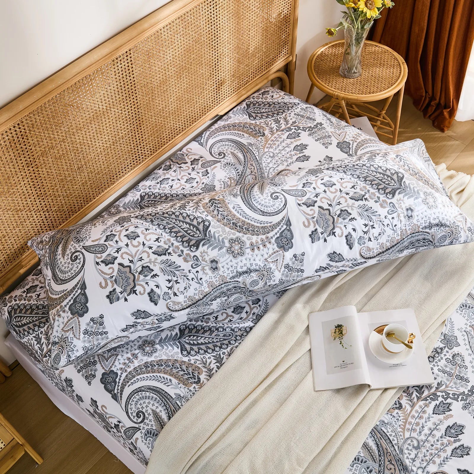 An elevated shot of a grey paisley cotton body pillowcase laid horizontally on a bed, with a book and teacup on the side.