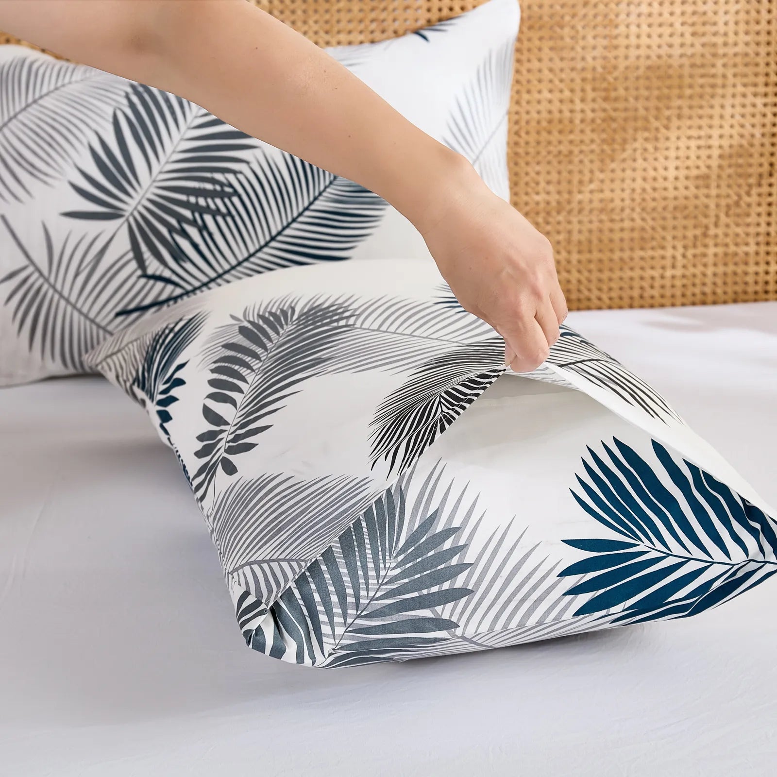 A hand holds and adjusts a cotton pillowcase featuring a modern palm leaf design, highlighting the soft texture and quality of the fabric.