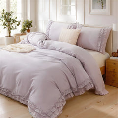 Lavender cotton double ruffle duvet cover on a wooden bed with pillows, a knit throw, and green plants near a window.