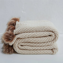 Folded light beige chunky knit throw blanket with brown faux fur pom-poms