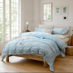 Light blue cotton ruffle bedding set on a bed with a small table and lamp by a large window.