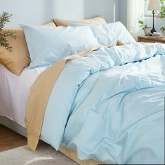 Angled top-down view of a light blue duvet with layered pillows and a black lamp on a wooden stand.