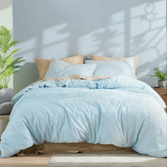 Front view of a light blue solid cotton duvet on a single bed with mixed pillows, green plants and window light shadows.