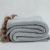 Folded light heather grey chunky knit throw blanket with brown faux fur pom-poms