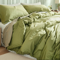 Asymmetric bedroom view with a matcha green duvet set between different-sized wooden tables and a yellow flower vase.