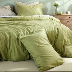 Matcha green duvet with casually stacked pillows beside a pink nightstand holding a vase of white flowers and green fruit.