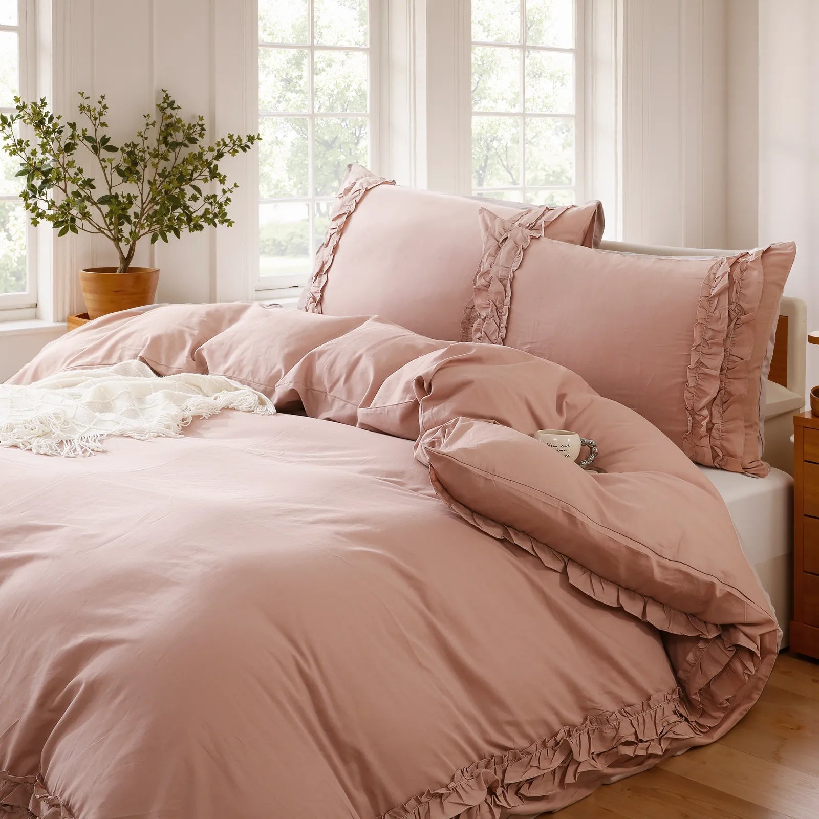Pink cotton double ruffle duvet cover and pillows on a bed beside a window with a green plant.
