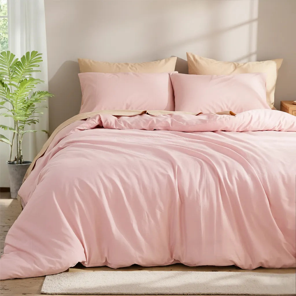 Corner view of a pink solid cotton duvet on a bed with four pillows, a green plant on the left and a wooden nightstand on the right.