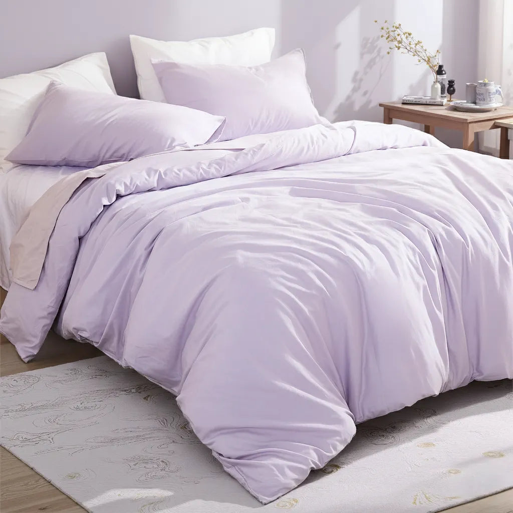 Layered purple and white pillows on a purple cotton duvet, with dried flowers in a vase and books on a wooden bedside table.