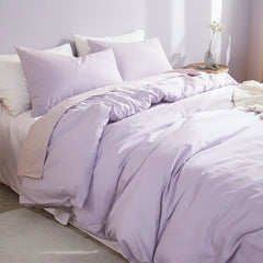 Softly textured purple cotton duvet on a bed with a round wooden nightstand holding white blooms in a clear vase.
