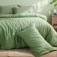 Sage green solid cotton duvet with color-block pillows, beside a nightstand with stacked books and a trailing plant.