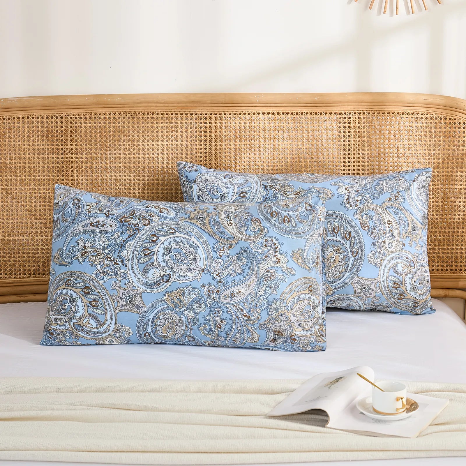 Two light blue cotton pillowcases with a detailed paisley pattern, stacked neatly on a bed with a rattan headboard.