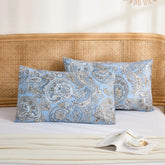 Two light blue cotton pillowcases with a detailed paisley pattern, stacked neatly on a bed with a rattan headboard.