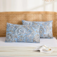 Two light blue cotton pillowcases with a detailed paisley pattern, stacked neatly on a bed with a rattan headboard.