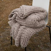 A cozy taupe chunky knit blanket is neatly folded on a chair outdoors, showing its soft, thick texture.